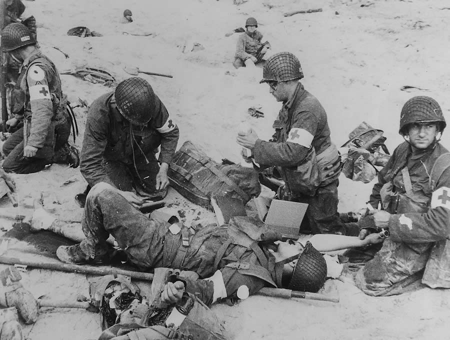 Original Caption: American Medics render first aid to troops in the initial landing on Utah Beach. Les Dunes de Madeleine, Northern Coast, France. In the background other members of the landing parties dig into the soft sand of the beach. June 6, 1944.